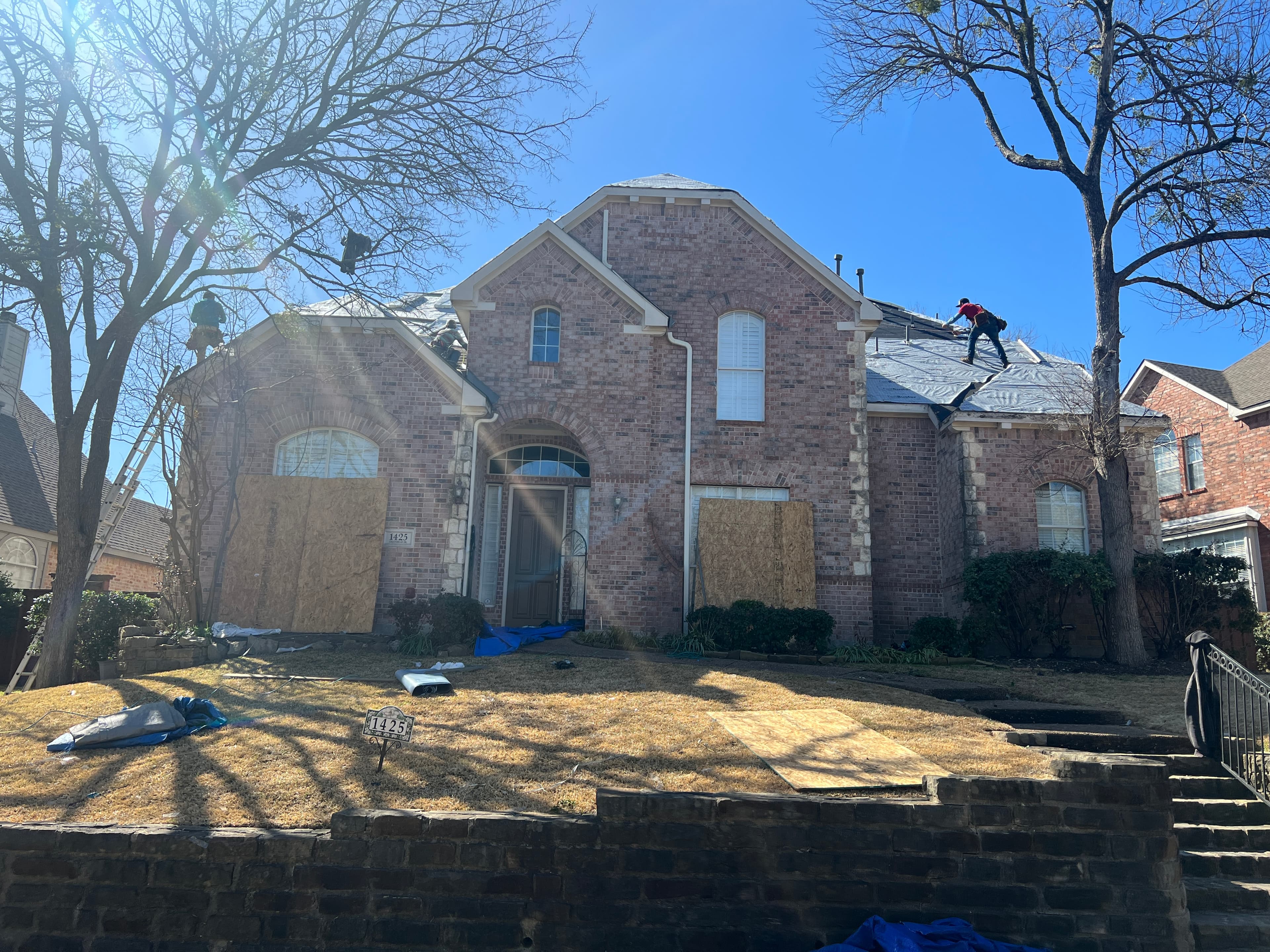 Front elevation of a two-story brick residence showing window plywood protection and synthetic underlayment installation on the roof.